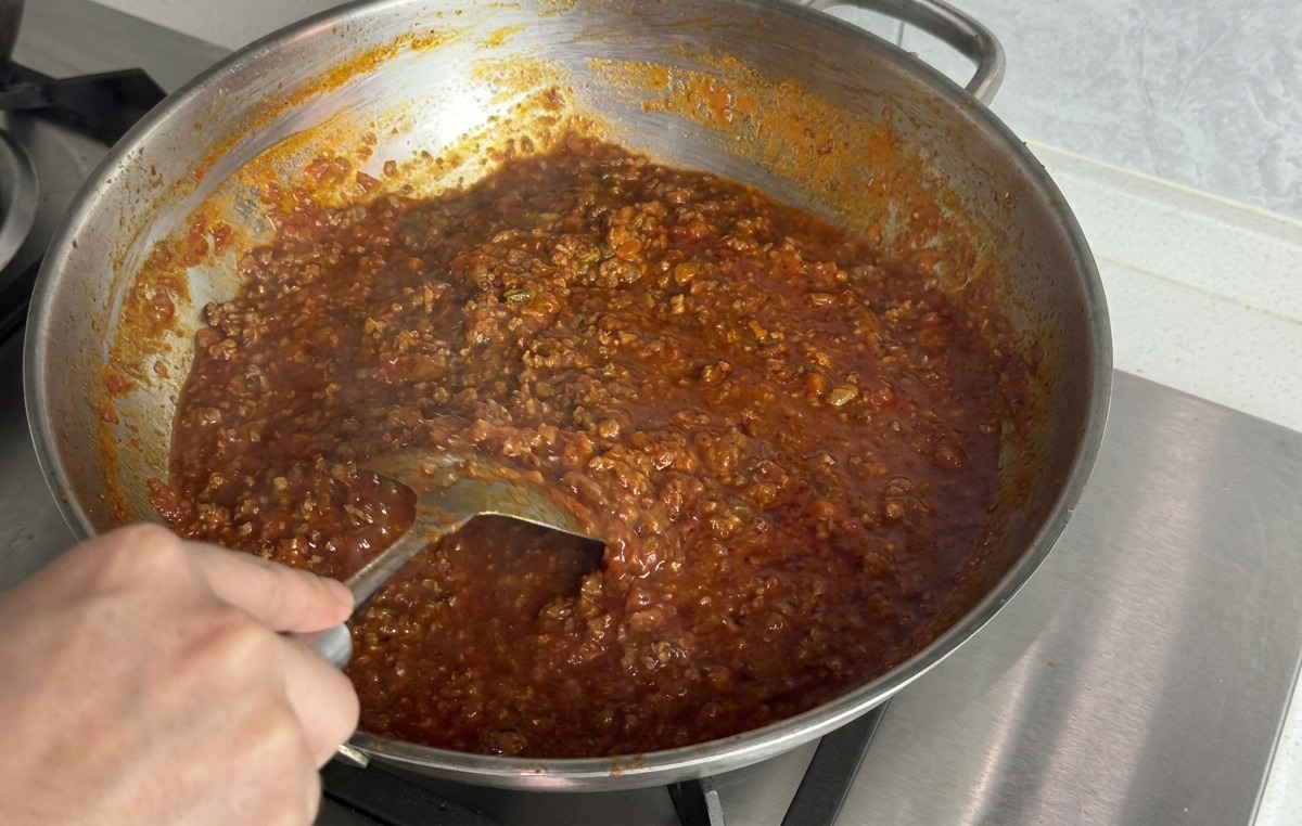 Una mano usa una spatola di metallo per incorporare un pezzetto di burro in un ragù di carne denso e rosso in una padella dacciaio.
