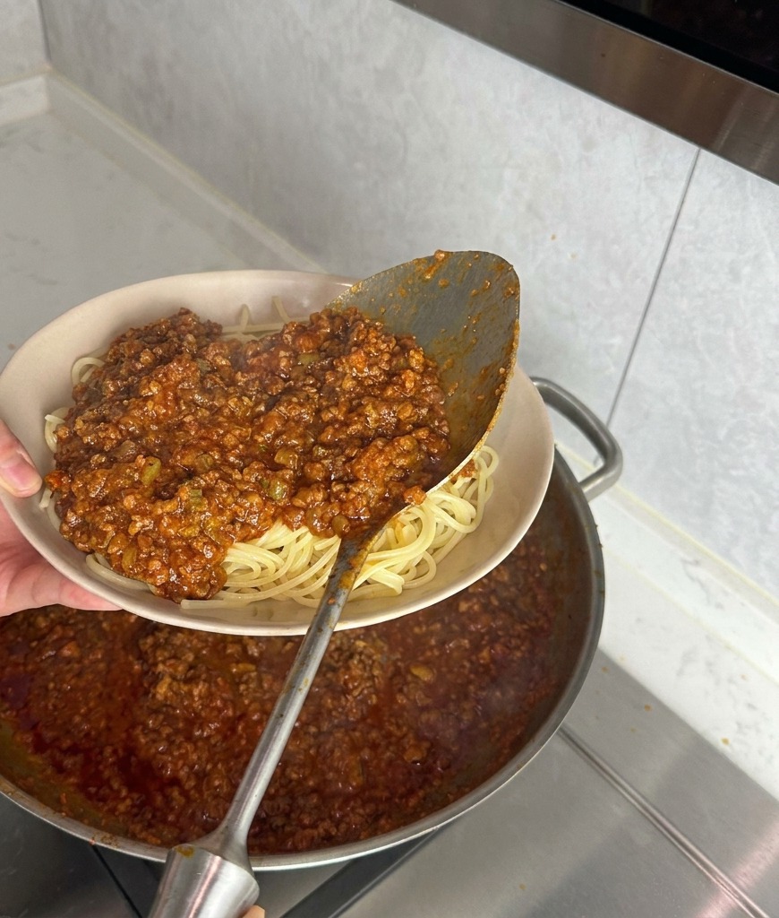 Un mestolo di metallo versa un sugo di carne denso e rosso su spaghetti bianchi cotti in una ciotola beige.
