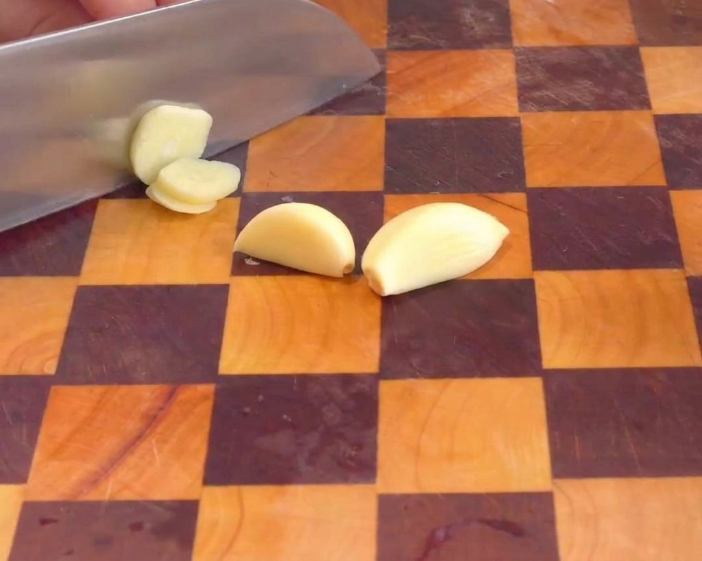 Una mano che usa un coltello da chef per tagliare gli spicchi daglio sbucciati a fettine sottili su un tagliere.