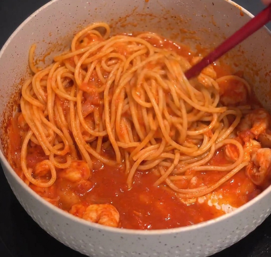 Spaghetti saltati con salsa rossa ai gamberi usando pinze da cucina in una pentola puntinata.