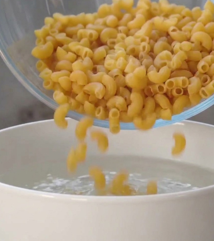 Fusilli crudi che cadono da una ciotola di vetro trasparente in una pentola bianca piena dacqua.