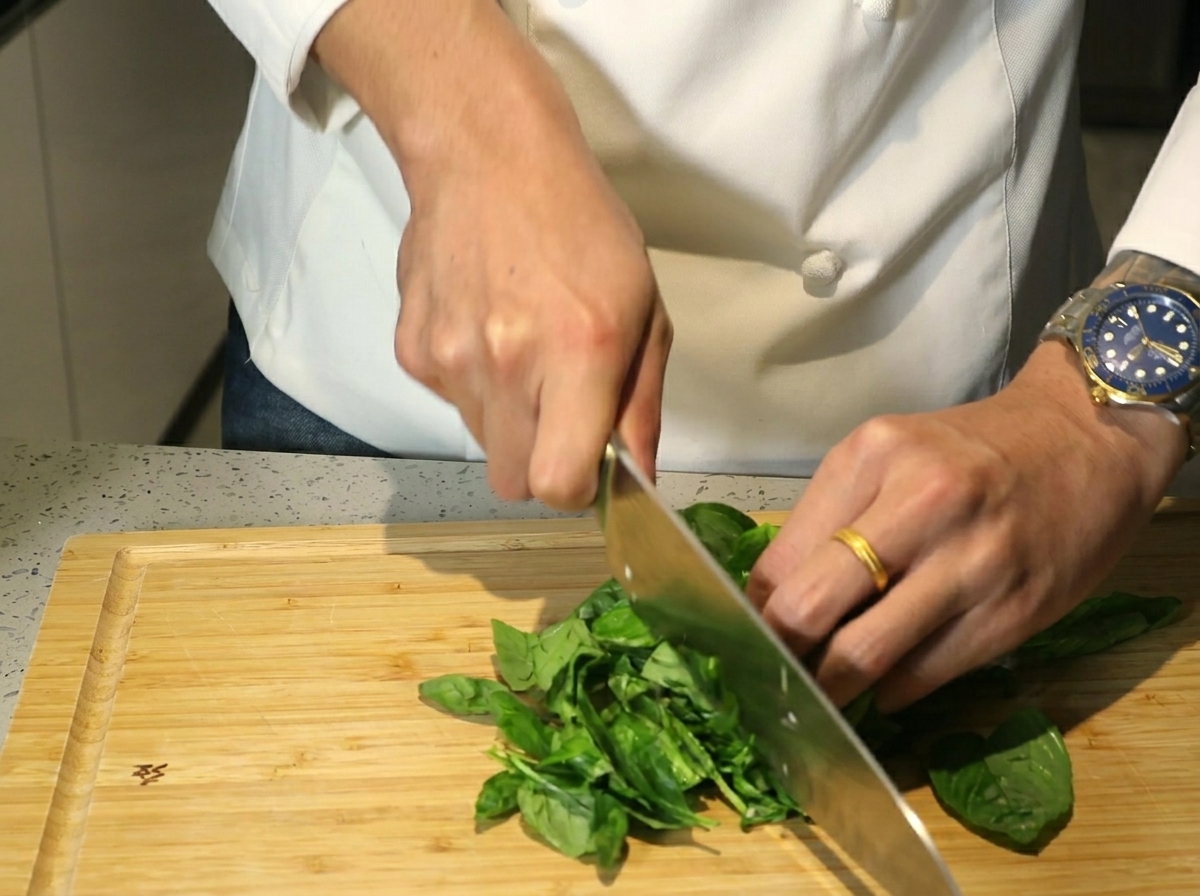 Mani che usano un grosso coltello per tritare una pila di foglie di basilico verde su un tagliere di legno.