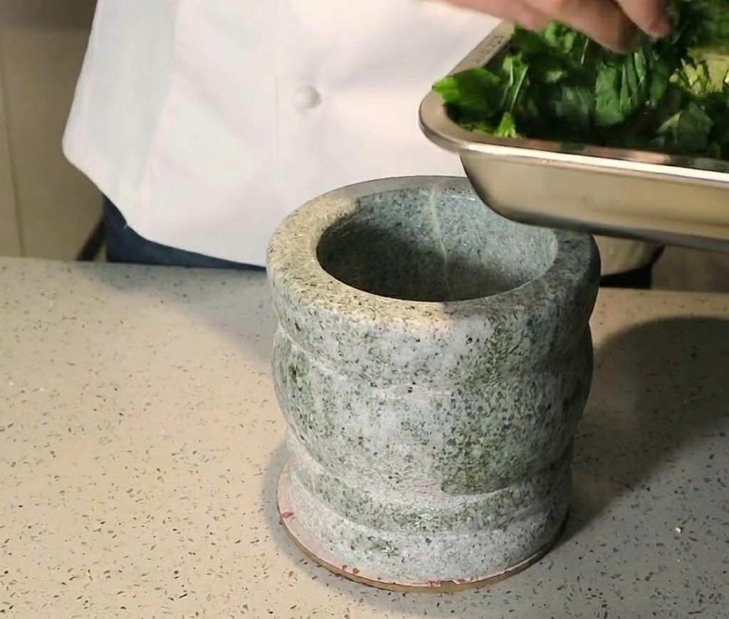 Una persona che aggiunge foglie di basilico fresco tritate in un mortaio di granito pesante.