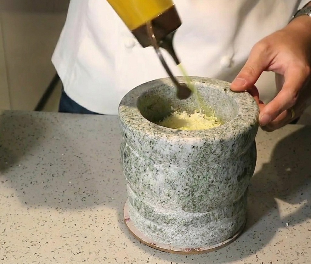 Olio doliva dorato versato da una bottiglia in un mortaio di pietra pieno di erbe e noci pestate.