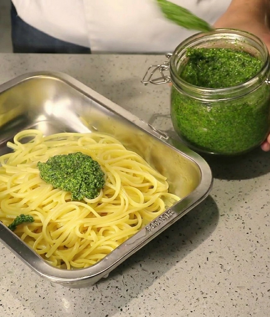 Densa salsa verde versata con un cucchiaio da un vasetto di vetro su spaghetti caldi in un vassoio dacciaio.