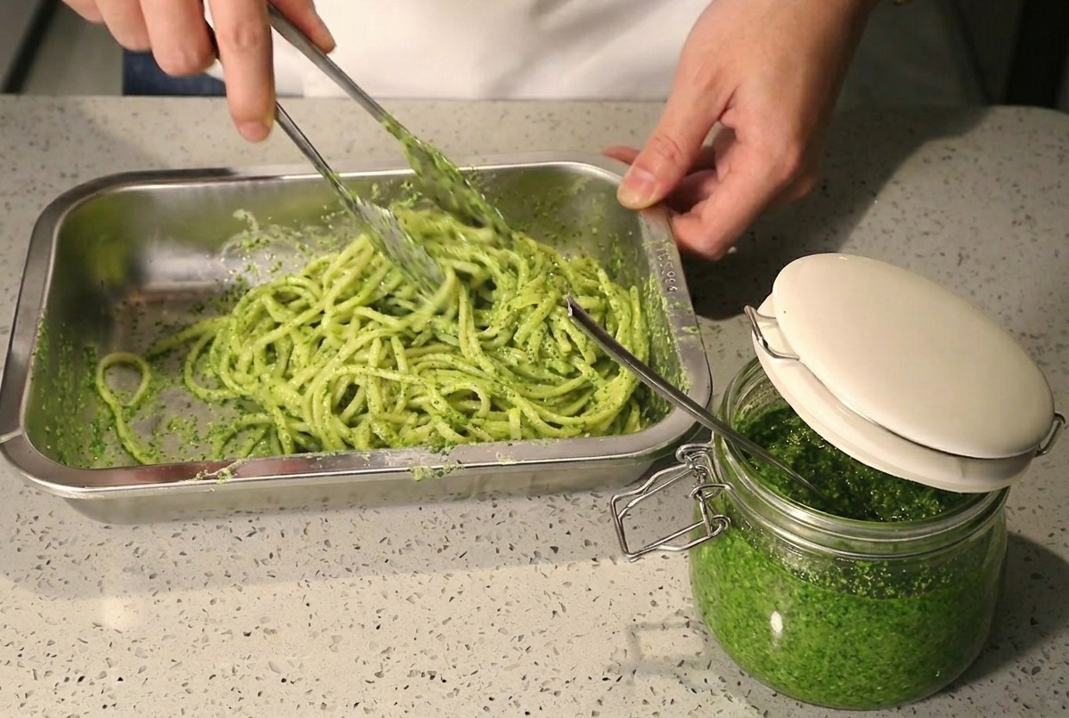 Uso di pinze metalliche per mescolare spaghetti con pesto verde e panna bianca in un vassoio.