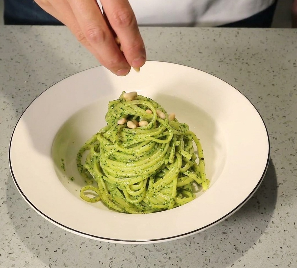 Una mano che sparge pinoli tostati dorati su una porzione di pasta al pesto impiattata in una ciotola bianca.