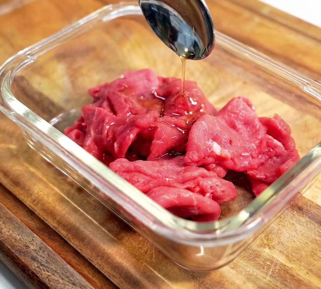 Un cucchiaio di metallo versa il condimento scuro sulle fettine di manzo crudo in una ciotola di vetro.
