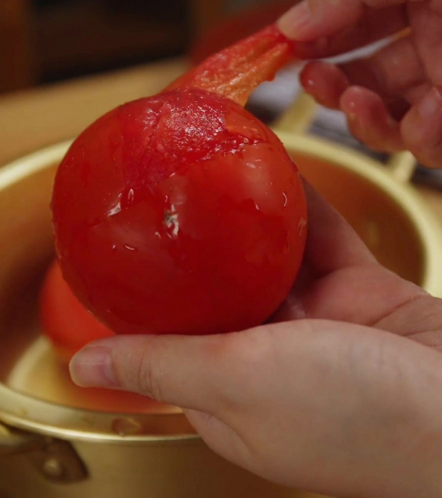 Primo piano di una mano che rimuove la buccia sottile e rossa da un pomodoro intero ammorbidito.
