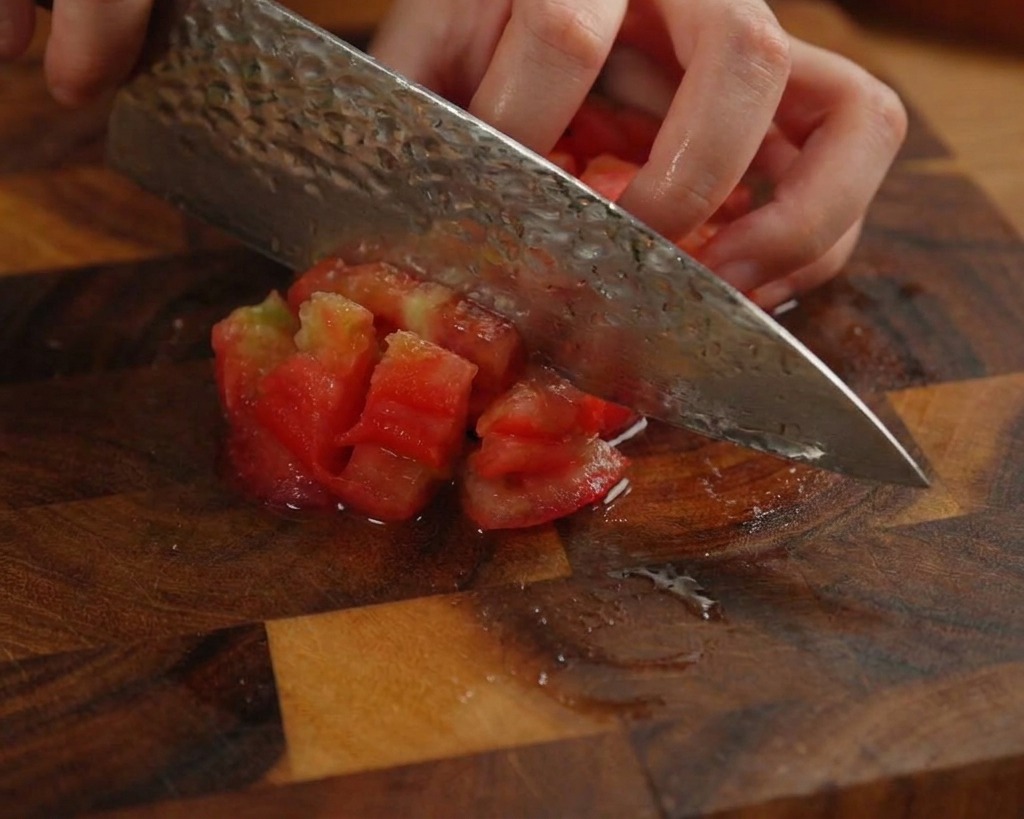 Una persona che taglia a cubetti un pomodoro pelato su un tagliere di legno scuro usando un coltello da cucina.