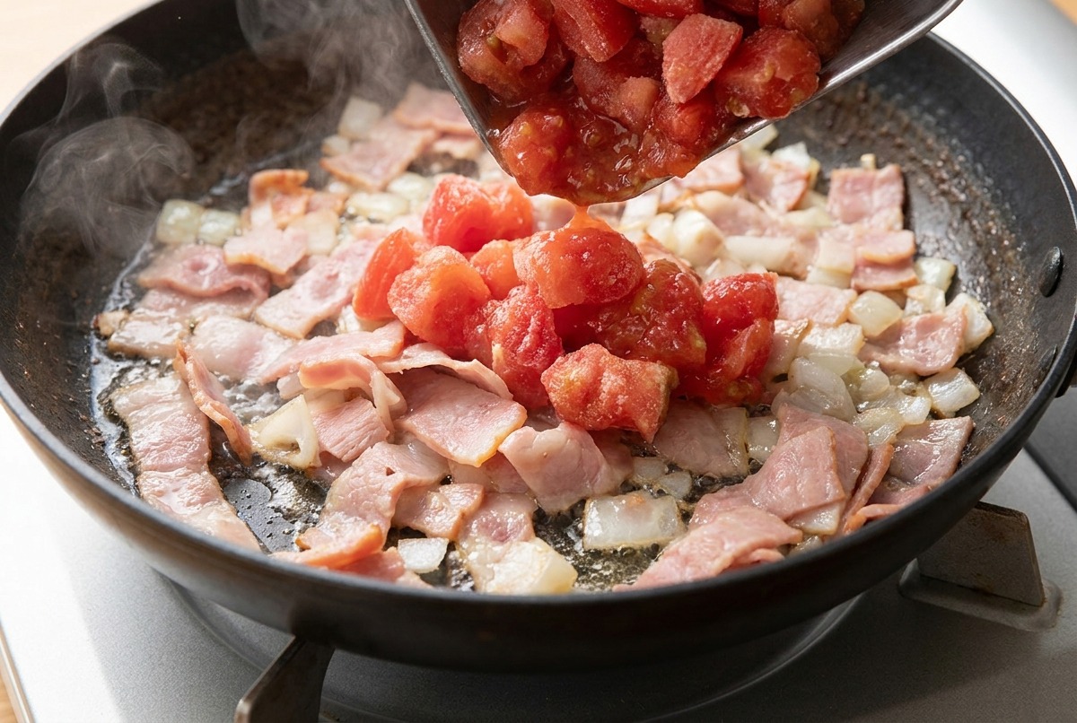 Bacon a fette e cipolle tritate in cottura in padella con laggiunta dei pomodori a cubetti.