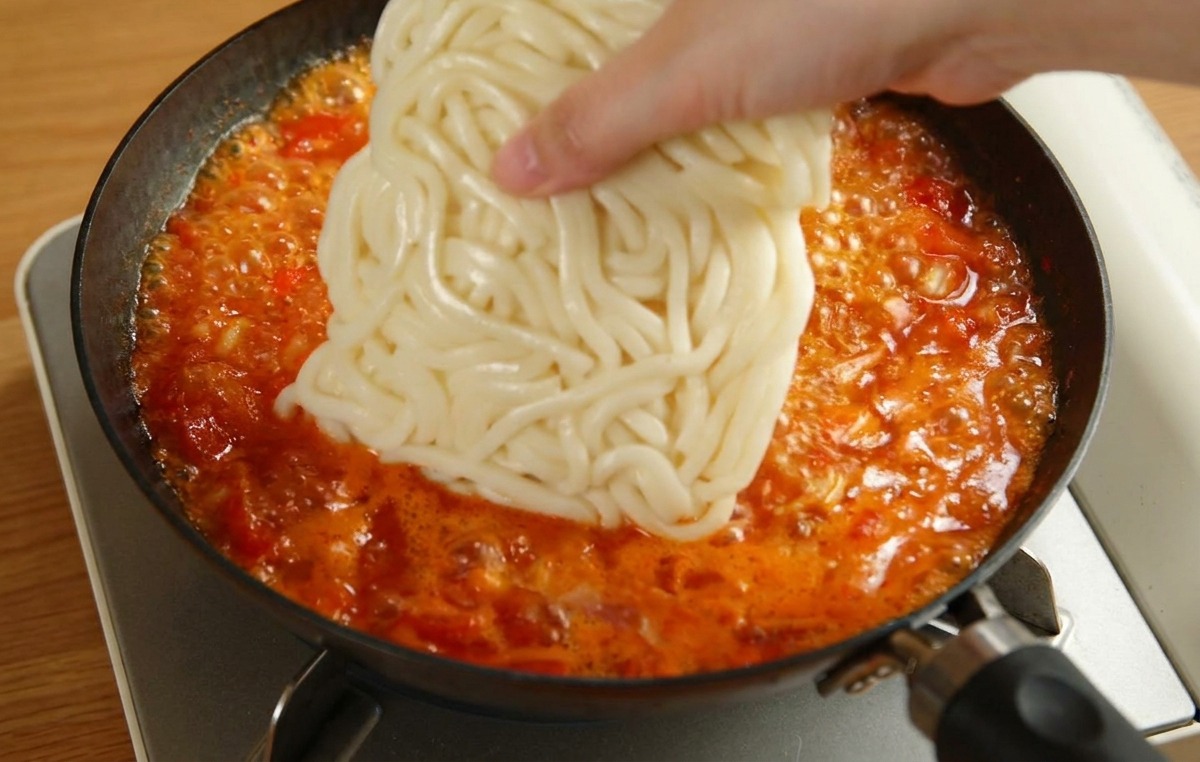 Una mano che adagia un blocco di udon bianchi in una padella con salsa di pomodoro rossa in ebollizione.