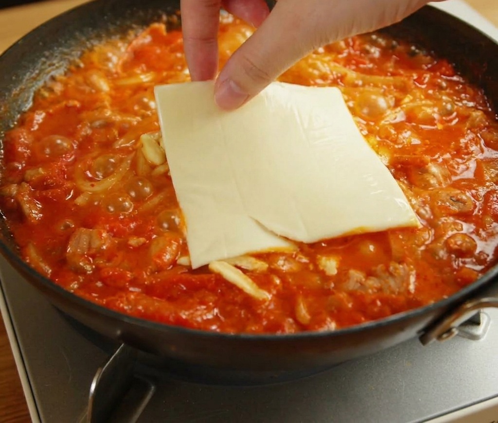 Una mano che posiziona una fetta quadrata di formaggio fuso giallo sopra la mozzarella e gli udon.