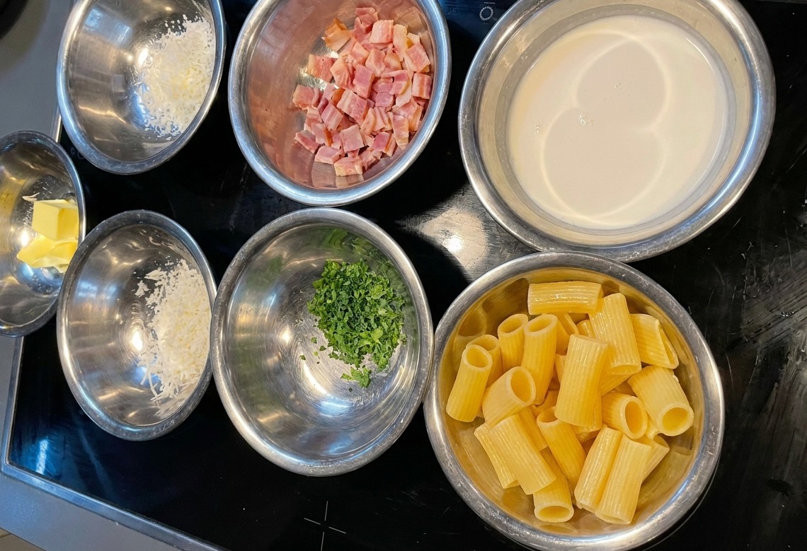 Diverse ciotole in acciaio inossidabile contenenti porzioni misurate di rigatoni, panna, bacon tritato, parmigiano e prezzemolo.