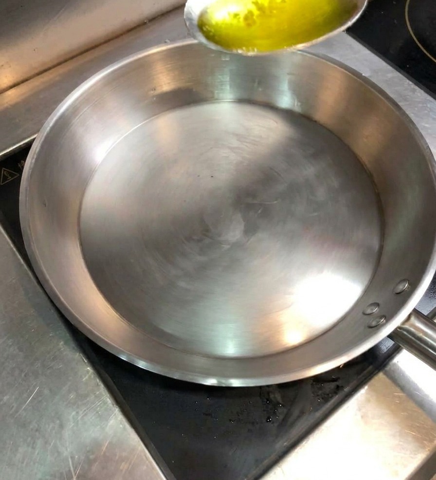 Un cucchiaio di metallo che versa olio in una padella di acciaio inossidabile pulita e calda.