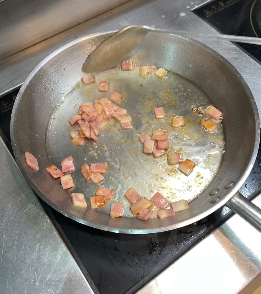 Pezzetti di bacon che sfrigolano e diventano dorati in padella, mossi con una spatola di metallo.