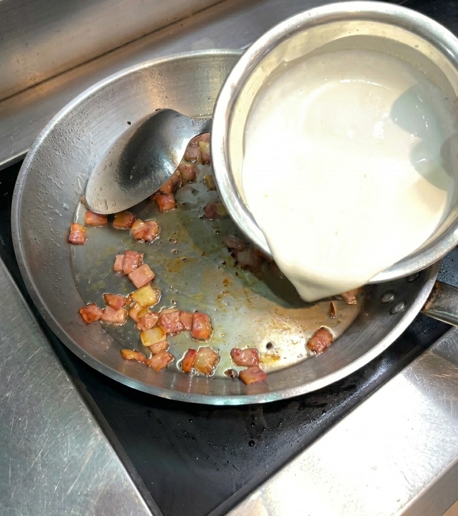 Panna densa versata da una ciotola in una padella con bacon croccante.