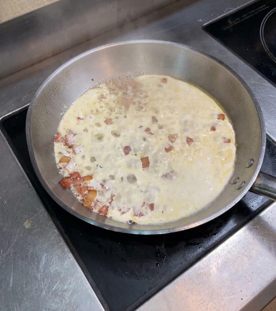 Miscela di panna e bacon che sobbolle in una padella larga sul fuoco.