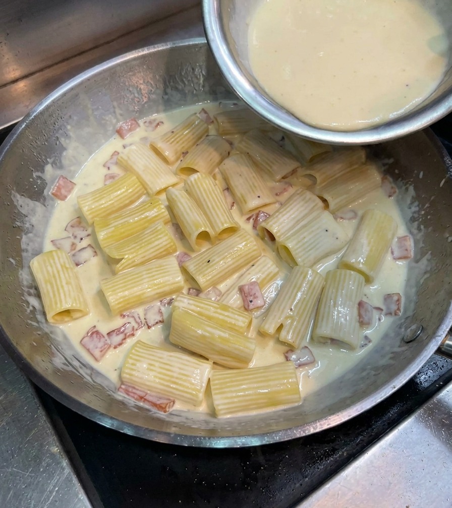 Composto giallo di uova e formaggio versato sopra i rigatoni in una grande padella di metallo.