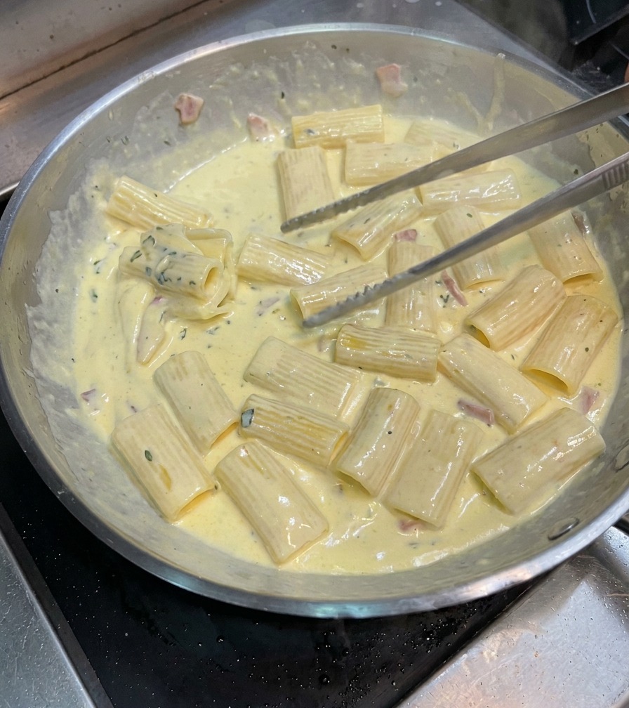 Pinze che saltano i rigatoni in padella per rivestirli di salsa carbonara densa.