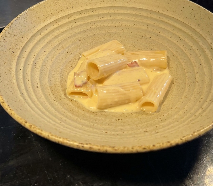 Porzione di rigatoni lucidi in salsa cremosa gialla con bacon, serviti in una ciotola di ceramica.