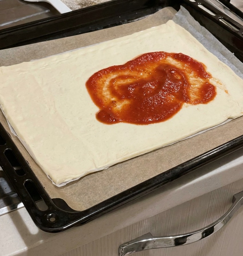 Salsa di pomodoro densa e rossa versata al centro di un impasto rettangolare crudo su una teglia.