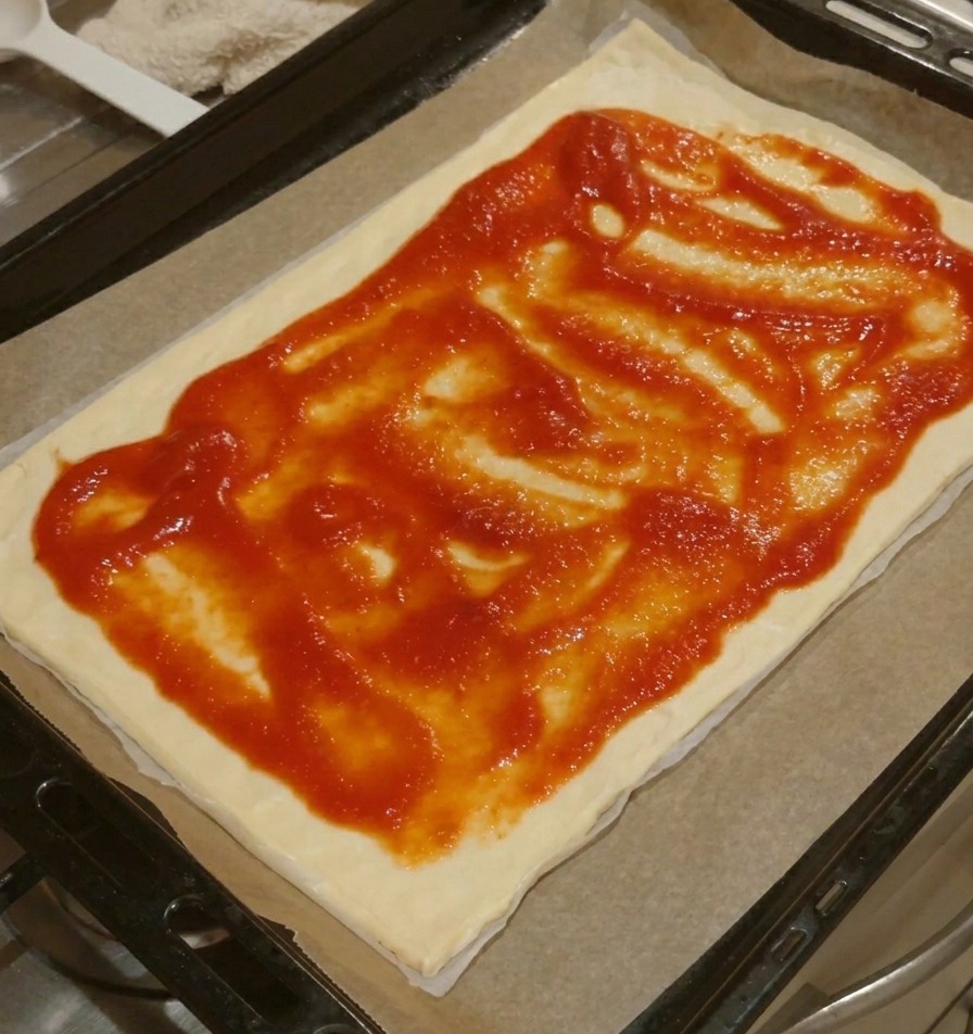 Una base per pizza rettangolare con salsa di pomodoro rossa stesa uniformemente sulla superficie, lasciando un bordo libero.