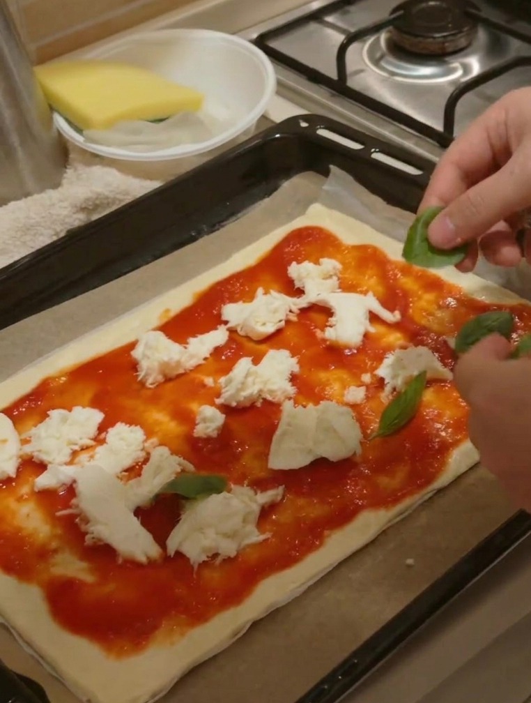 Mani che posizionano foglie di basilico verde fresco su una pizza già condita con salsa e pezzi di formaggio.