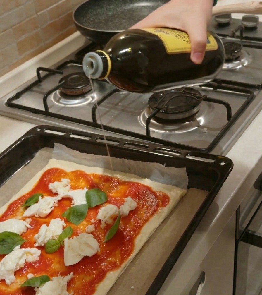 Una mano che versa un filo dolio doliva da una bottiglia di vetro su una pizza cruda condita con pomodoro, mozzarella e basilico.