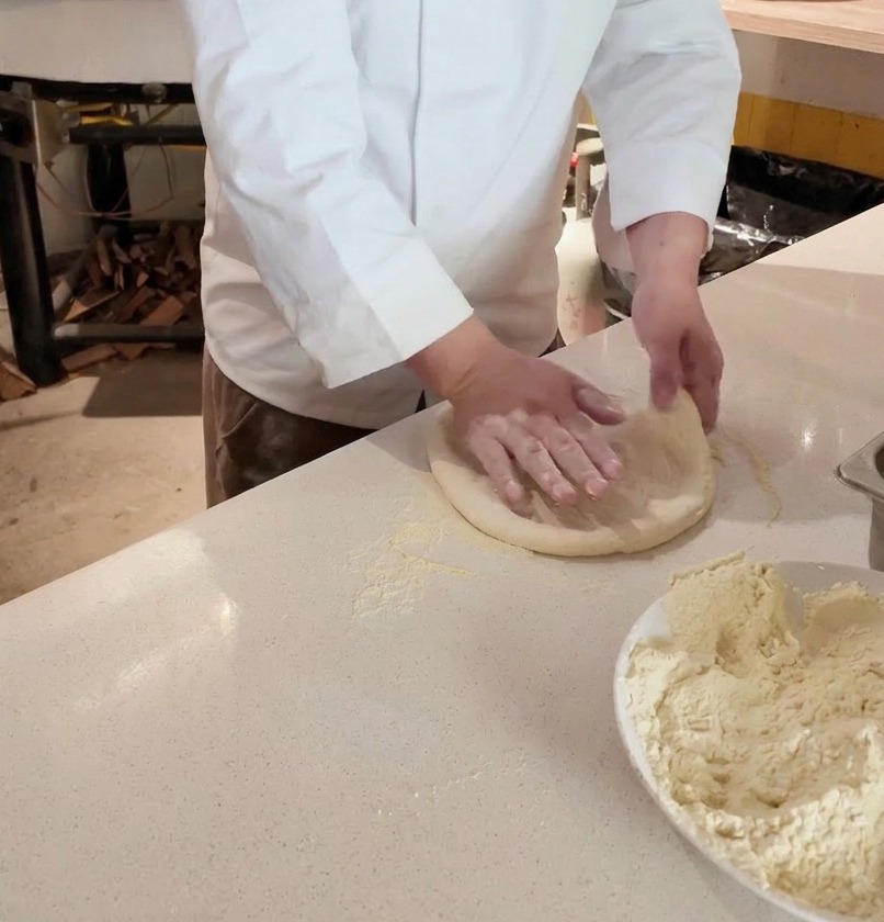 Mani di uno chef che stendono un panetto di pizza su un piano infarinato davanti a un forno a legna.