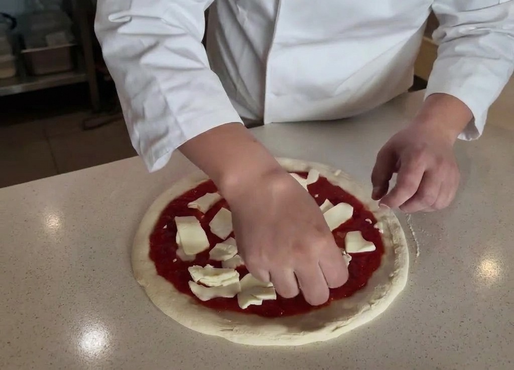 Mani che distribuiscono pezzi di mozzarella di bufala bianca su una base condita con salsa di pomodoro.