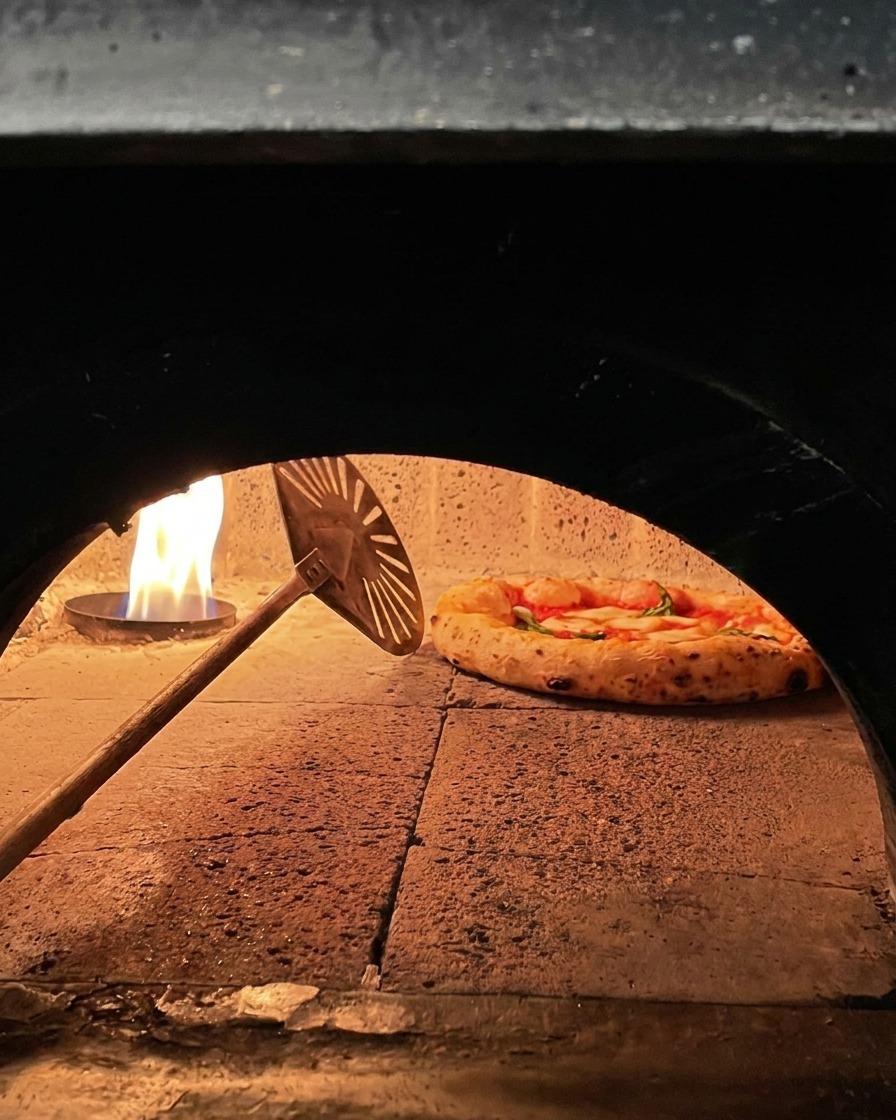 Pizza Margherita in cottura in un forno a legna mentre viene ruotata con un palino metallico vicino alla fiamma.