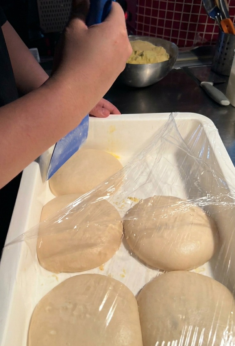 Una persona usa una spatola di plastica blu per sollevare con cura un panetto di pizza rotondo da un contenitore bianco.