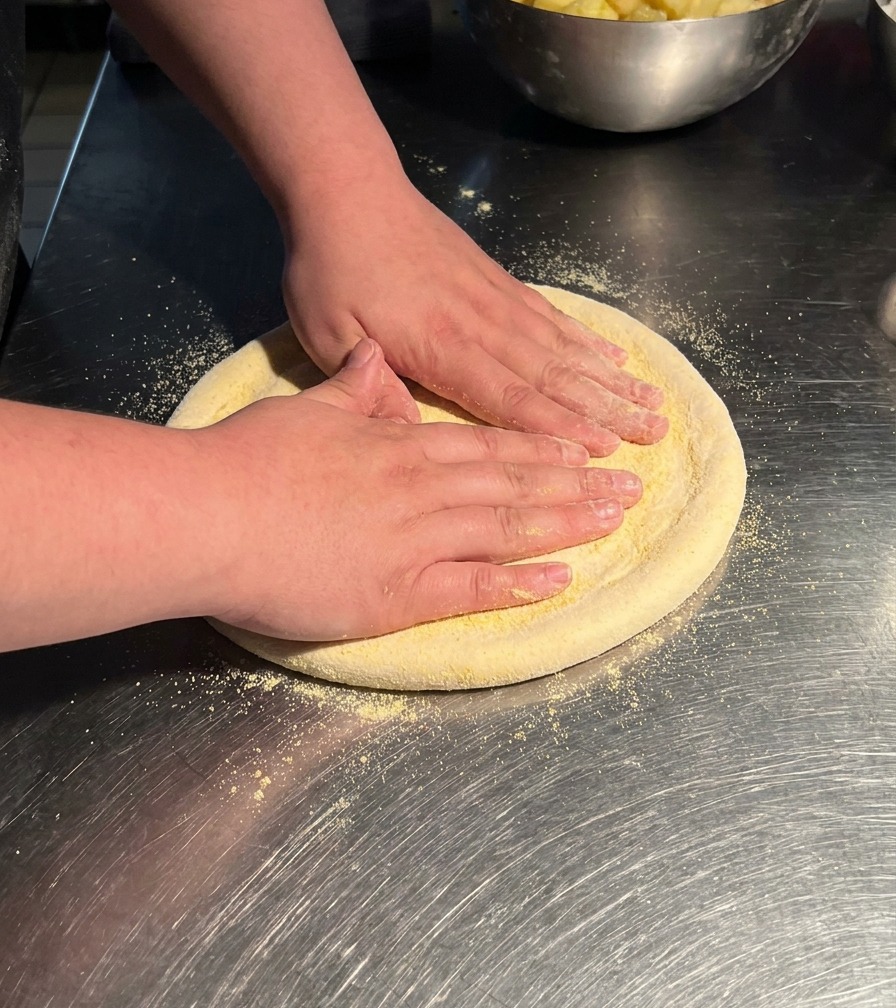 Mani che premono su un pezzo circolare di impasto per pizza su un tavolo in acciaio, stendendolo in un disco più grande.