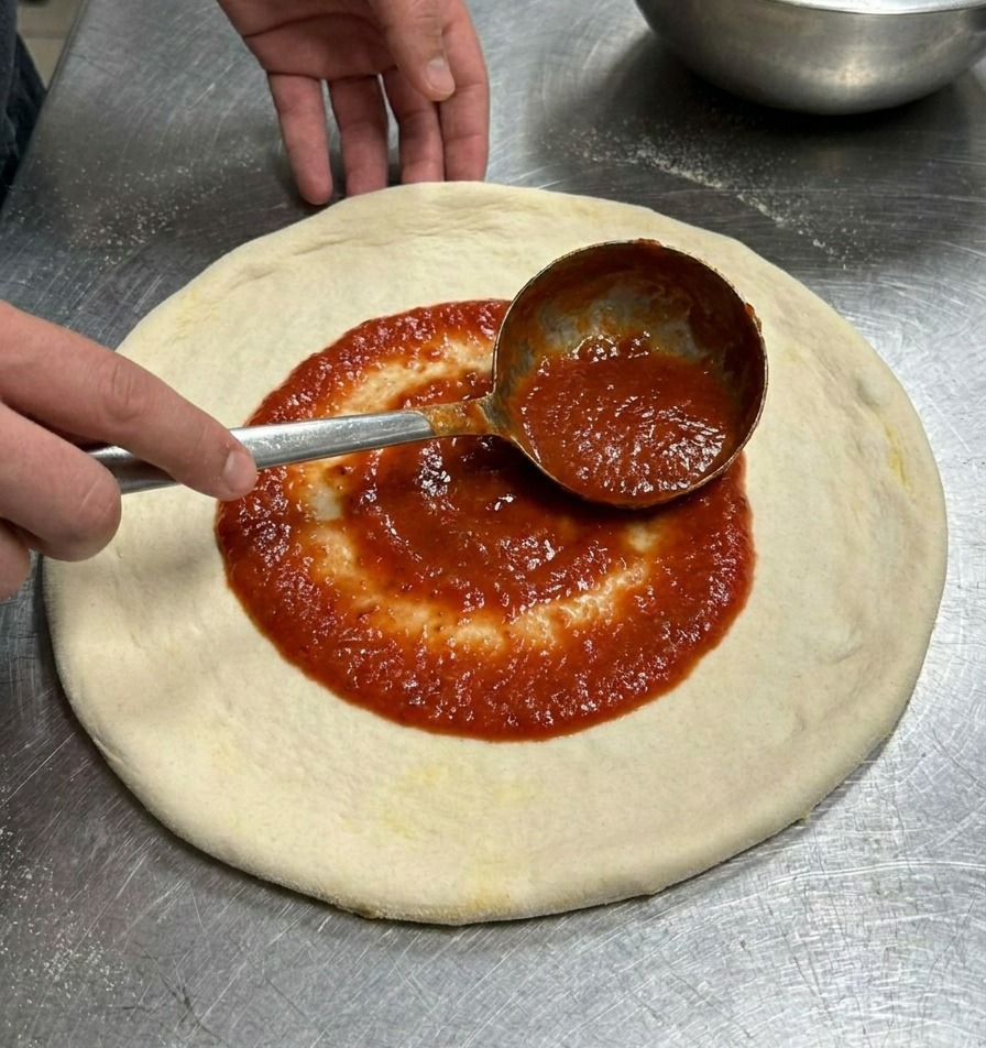 Un mestolo di metallo che stende la salsa di pomodoro rossa con un movimento circolare su un disco di pasta.