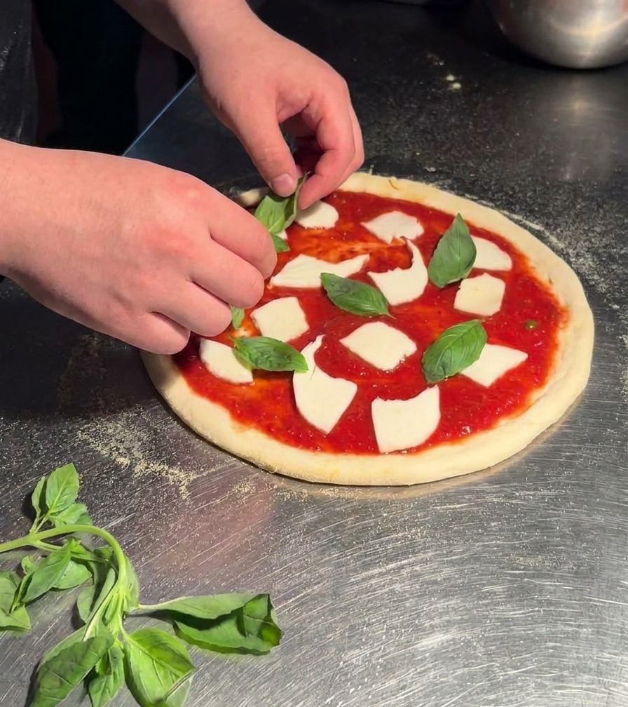 Mani che posizionano foglie di basilico verde brillante su una pizza Margherita cruda condita con pomodoro e mozzarella.