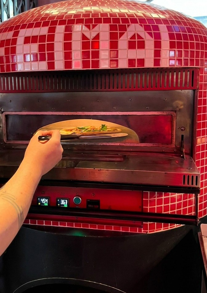 Una persona che fa scivolare una pizza cruda dalla pala allinterno di un forno tradizionale con piastrelle rosse.
