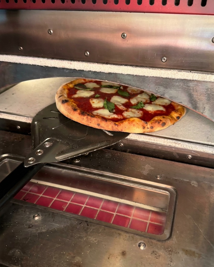Una pizza appena sfornata con i bordi del cornicione maculati, prelevata con una pala di metallo.