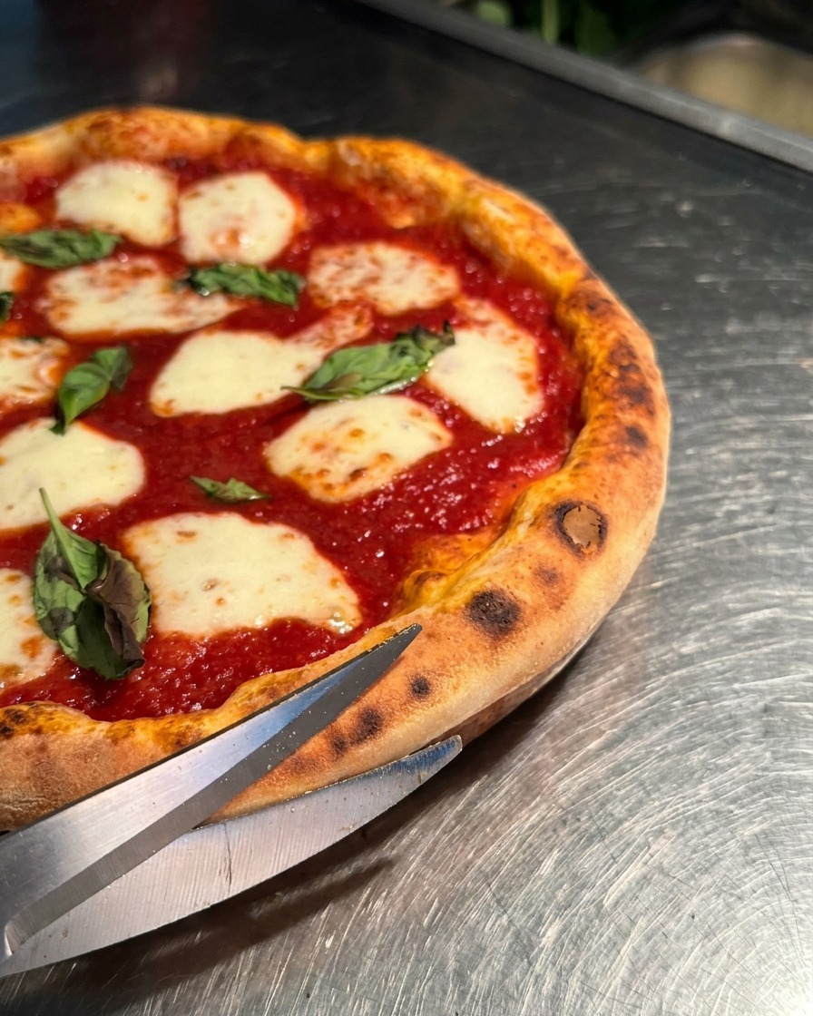 Primo piano di forbici da cucina affilate che tagliano il cornicione gonfio di una pizza Margherita calda.