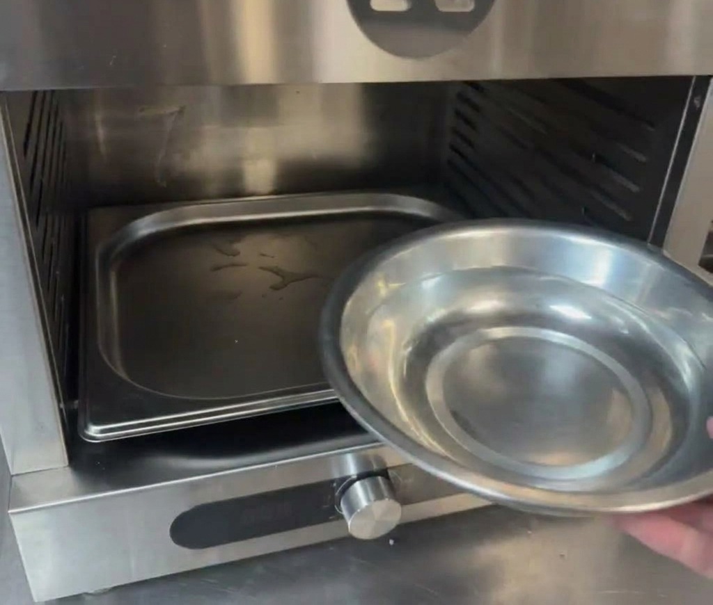 Una mano che versa acqua da una ciotolina di metallo in una teglia sul fondo della camera di un forno professionale.
