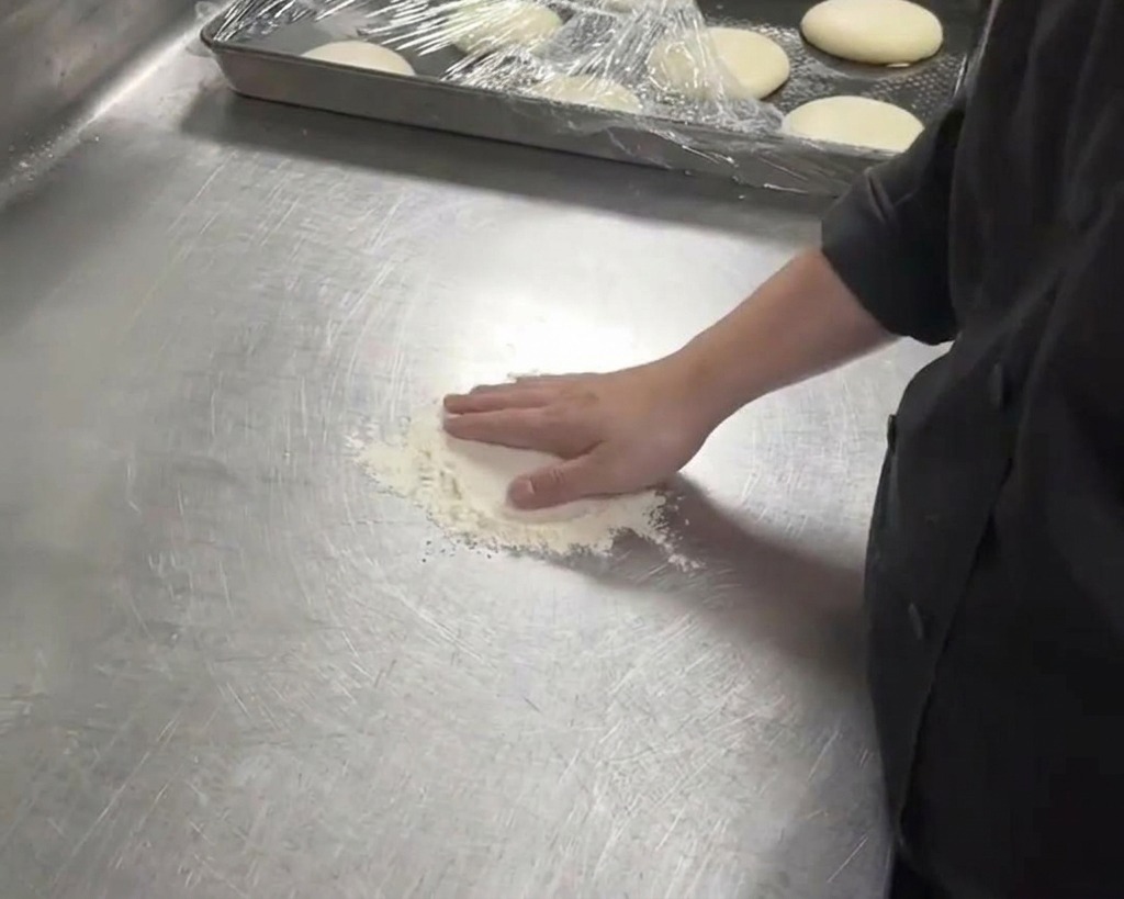 Un pizzaiolo con giacca nera che preme la mano su un panetto di pasta bianco su un bancone di metallo infarinato.