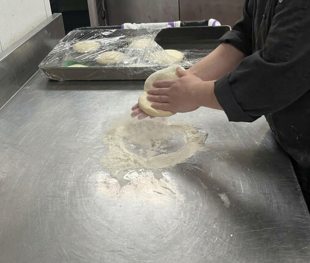 Mani che stendono delicatamente limpasto della pizza su una superficie infarinata, allargandolo in un cerchio.