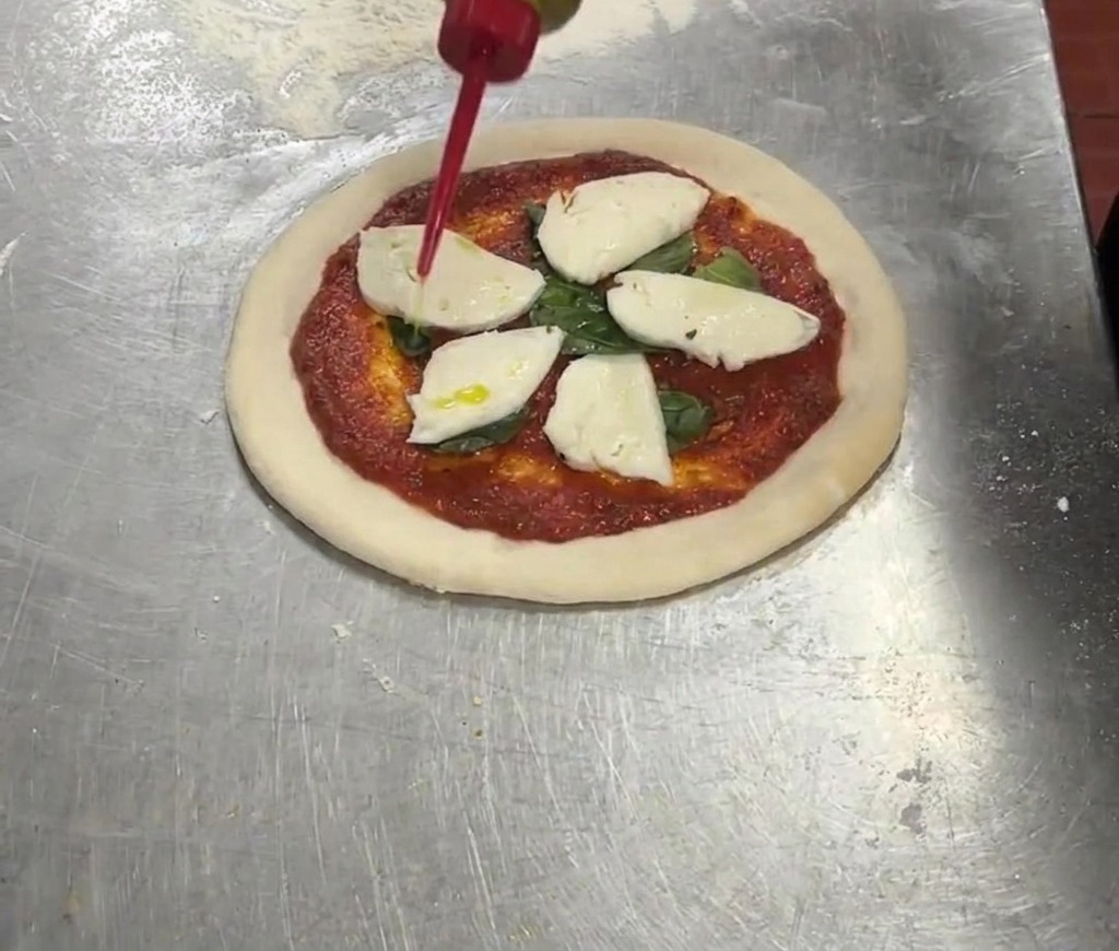 Un dosatore rosso che versa olio extravergine doliva a filo su una pizza guarnita con pomodoro, basilico e mozzarella.