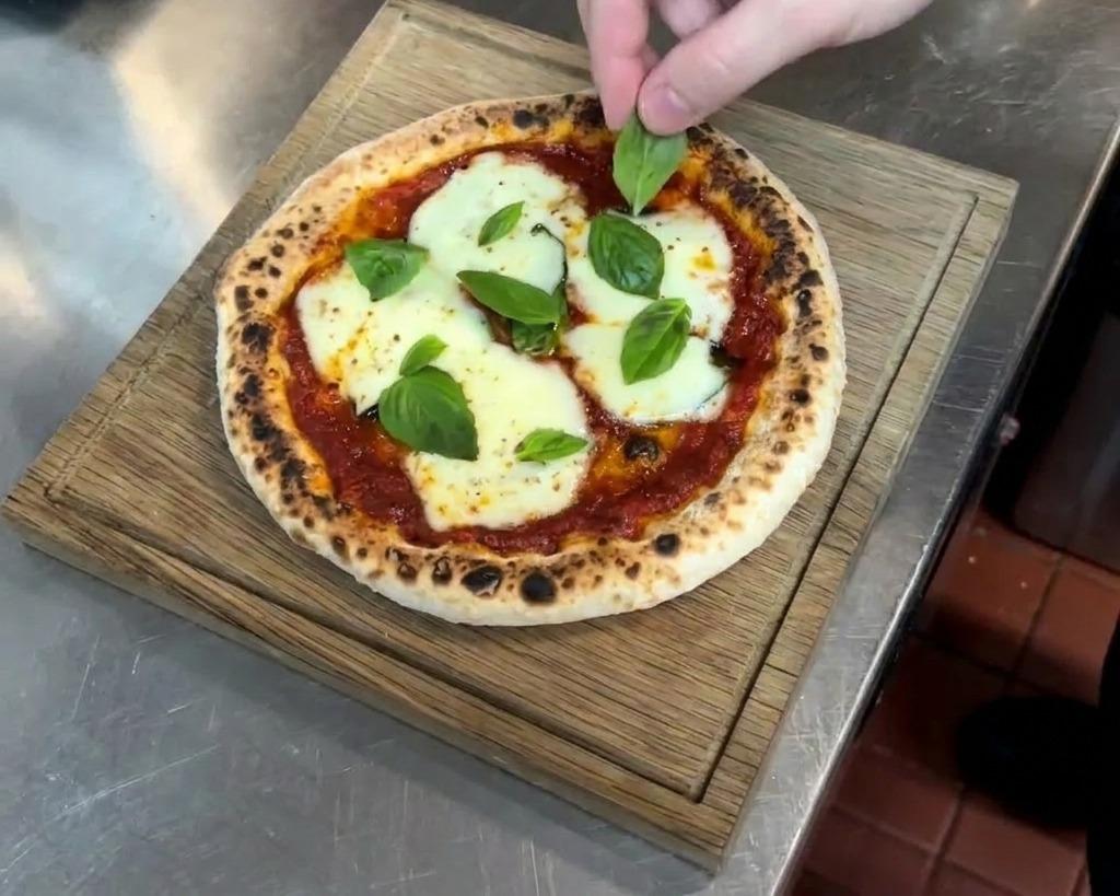 Mano che posiziona foglie di basilico su una pizza calda appena sfornata con mozzarella fusa e cornicione maculato.