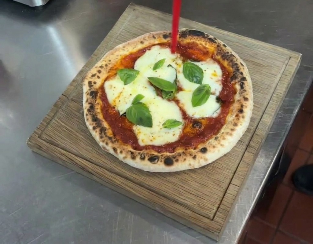 Un filo di olio extravergine doliva versato da un dosatore su una pizza Margherita finita appoggiata su un tagliere di legno.