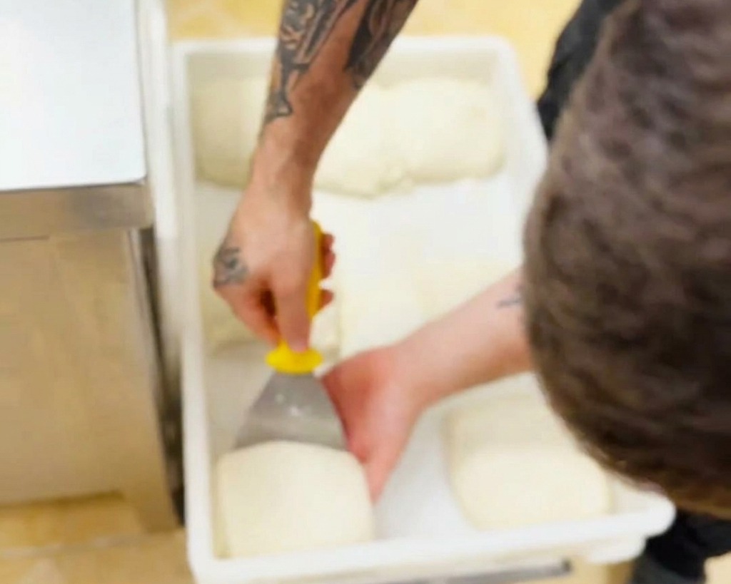 Una mano che usa un raschietto giallo per sollevare delicatamente un panetto di impasto bianco da un contenitore di plastica.