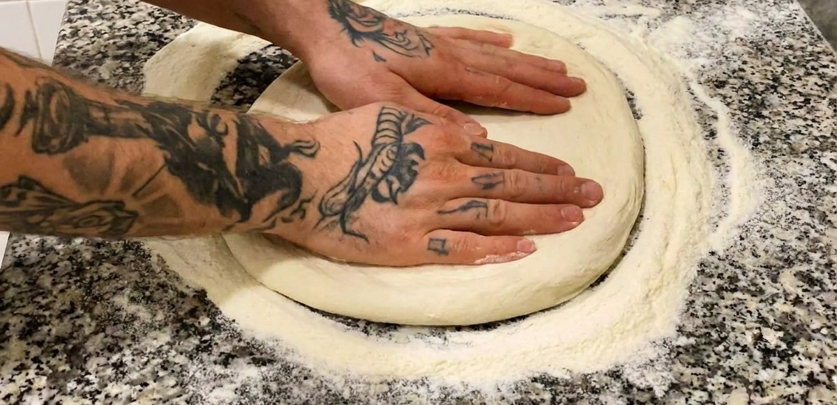Due mani tatuate che premono su un panetto di pizza infarinato su un piano di marmo grigio.