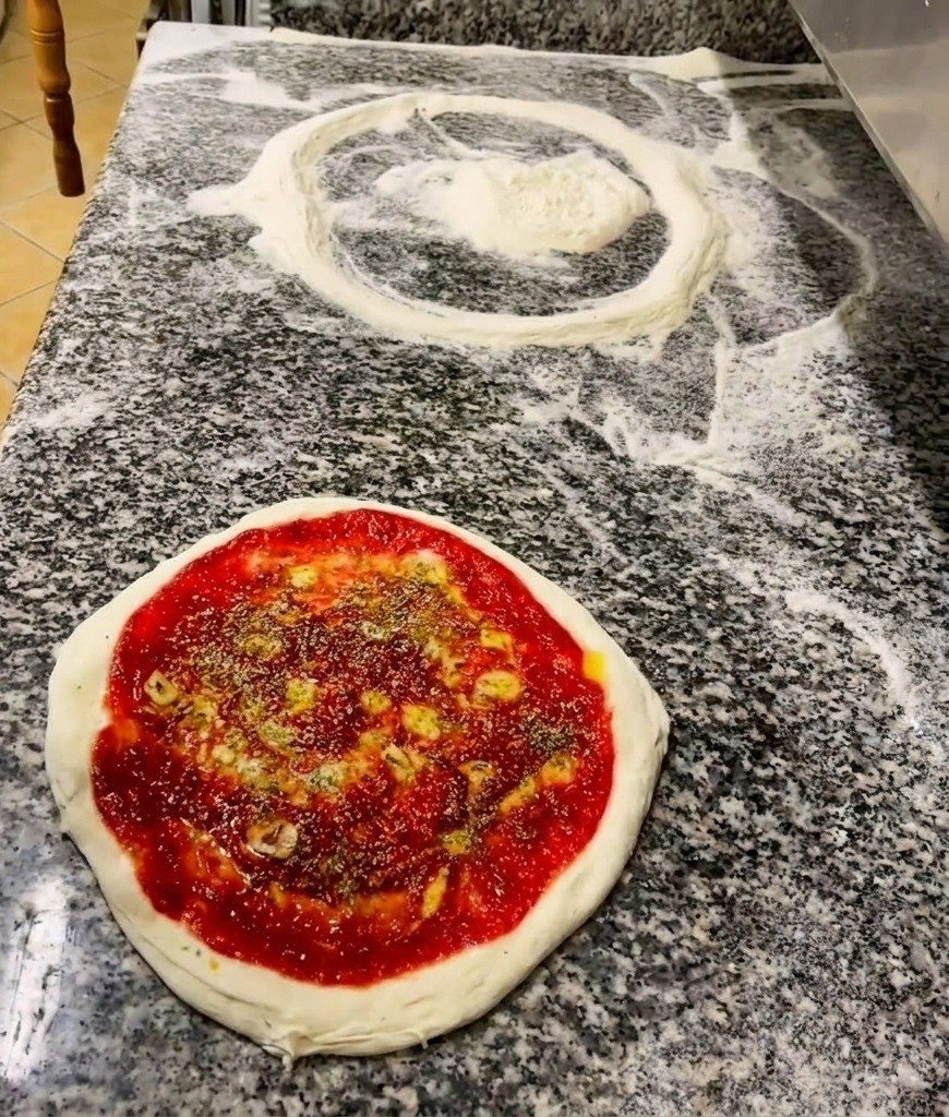 Una mano che sparge origano secco su una base pizza già condita con pomodoro e un filo dolio.