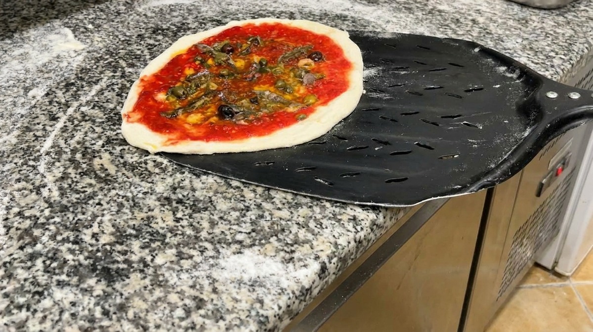 Una pizza pronta per essere infornata, adagiata su una pala di metallo nero forata.