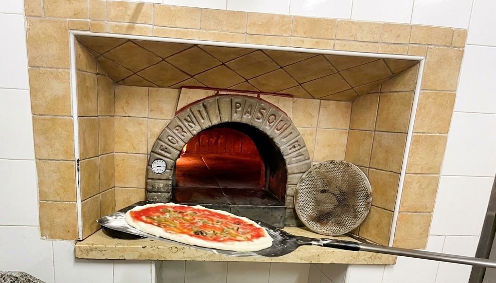 Una pizza su una pala dal manico lungo mentre viene inserita in un forno a legna tradizionale con fiamme vive.